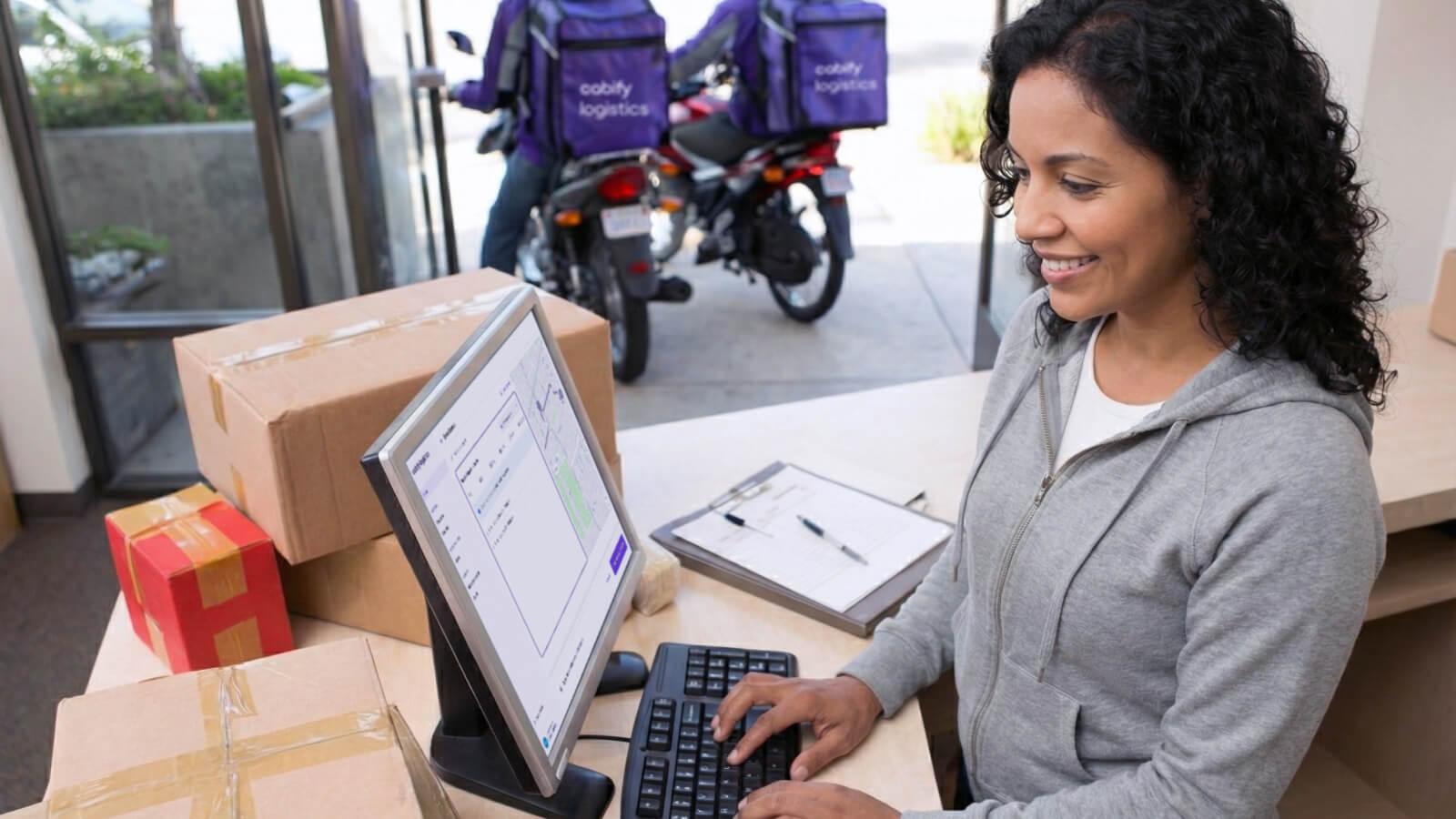 Mujer dueña de un negocio termina de actualizar estado de los envíos de sus productos, mientras dos conductores de Cabify Logistics se preparan a salir a hacer sus envíos.